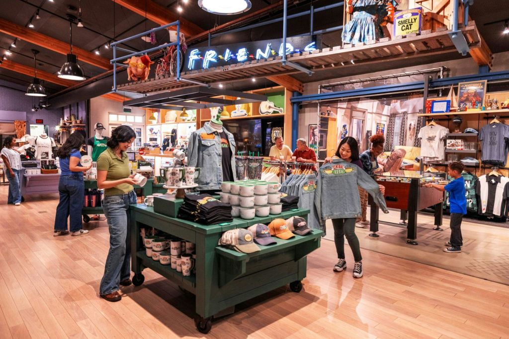 한 사람이 "Friends" 상품이 가득한 매장에서 "Friends" 테마의 스웨트셔츠를 들고 있습니다. 어린이들은 백그라운드에서 아이템을 탐색합니다.