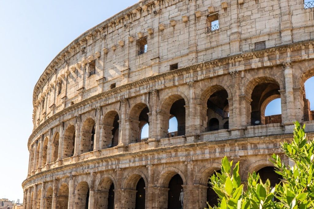 맑고 푸른 하늘 아래 여러 층과 창문이 있는 아치형 외관을 보여주는 로마의 콜로세움. 앞에 녹색 식물이 있습니다.