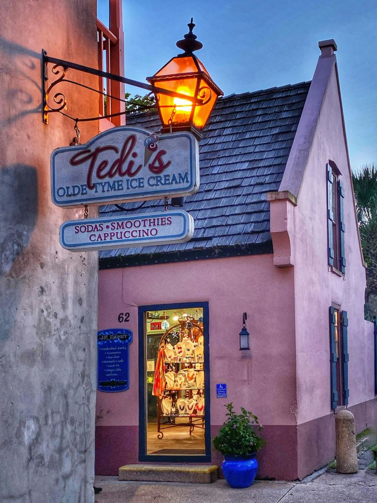 'Tedi's Olde Tyme Ice Cream' 이라는 간판이 켜져 있고 따뜻하게 빛나는 내부가 보이는 독특한 핑크 아이스크림 가게입니다.