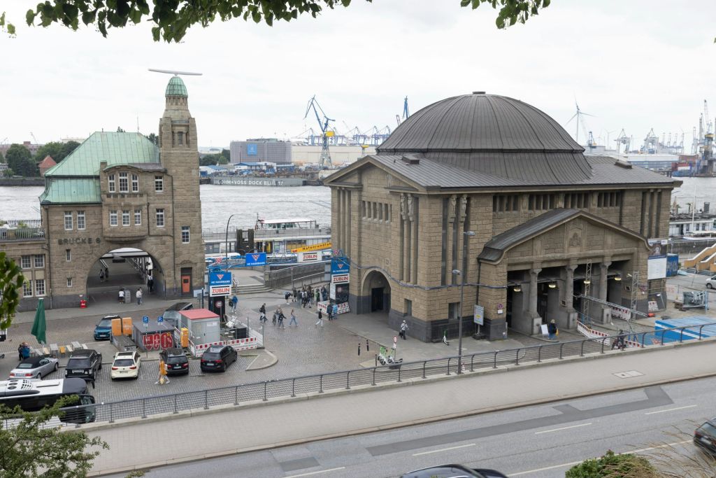 해안가의 커다란 석조 건물을 배경으로 크레인과 산업 구조물이 있는 도심 풍경. 사람과 차량이 보입니다.