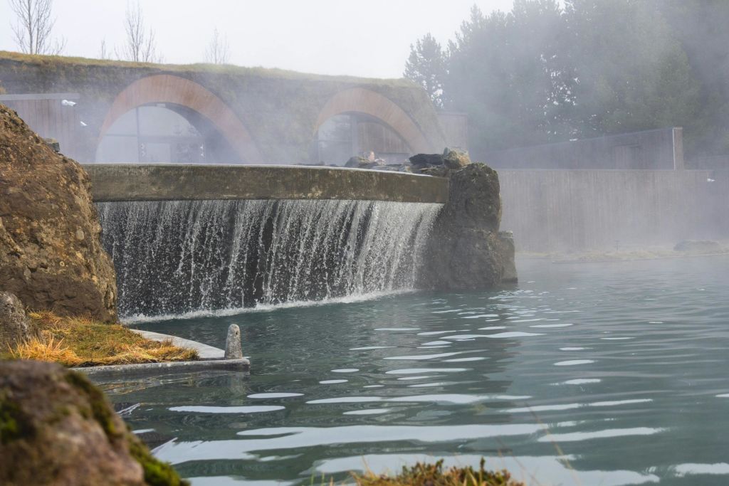 작은 폭포가 고요한 푸른 수영장으로 흘러들어가고, 안개 낀 하늘 아래 증기가 솟아오르고 아치형 다리가 배경으로 펼쳐집니다.
