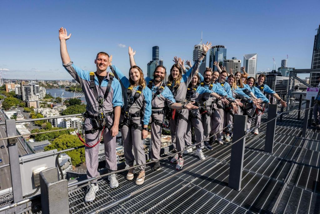 높은 건물과 강을 배경으로 한 도시의 옥상에서 안전장치를 착용한 사람들이 손을 흔들고 있습니다.