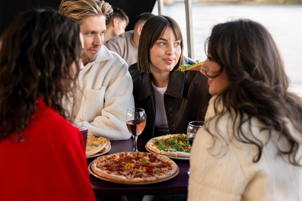 4명이 테이블에 앉아 다양한 피자와 와인잔을 마시며 미소를 짓고 대화에 참여합니다.