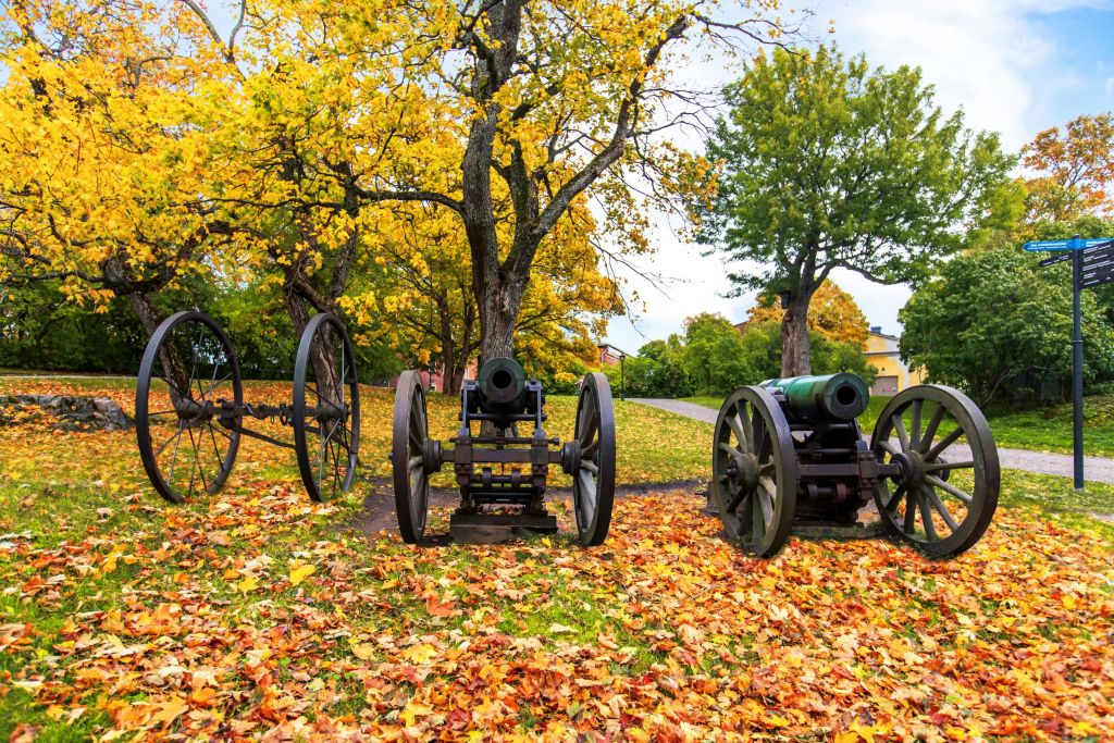 가을 나뭇잎으로 뒤덮인 잔디밭에는 바퀴 달린 오래된 대포 세 개가 전시되어 있으며, 나무를 배경으로 다채로운 풍경이 펼쳐집니다.