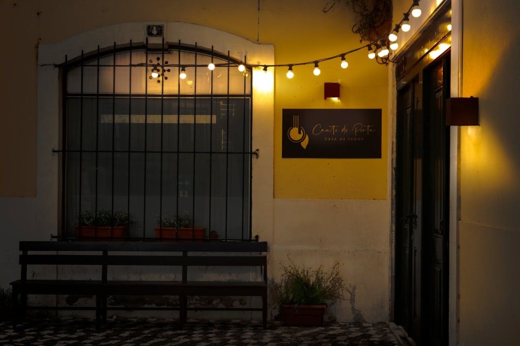 해질 무렵의 야외 카페 풍경에는 스트링 조명과 "Café de Praça" 라고 적힌 간판이 있습니다. 창문 아래에 벤치와 화분이 있습니다.