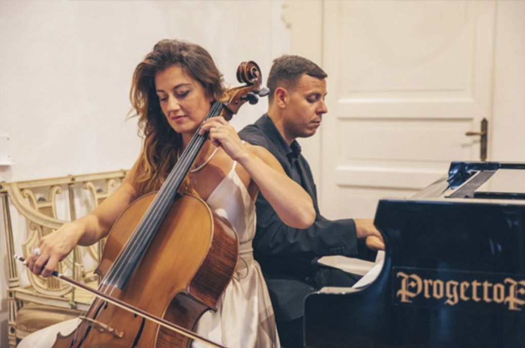 흰색 드레스를 입은 여성이 실내에서 피아노를 치는 검은색 셔츠를 입은 남성 옆에서 첼로를 연주하고 있습니다.