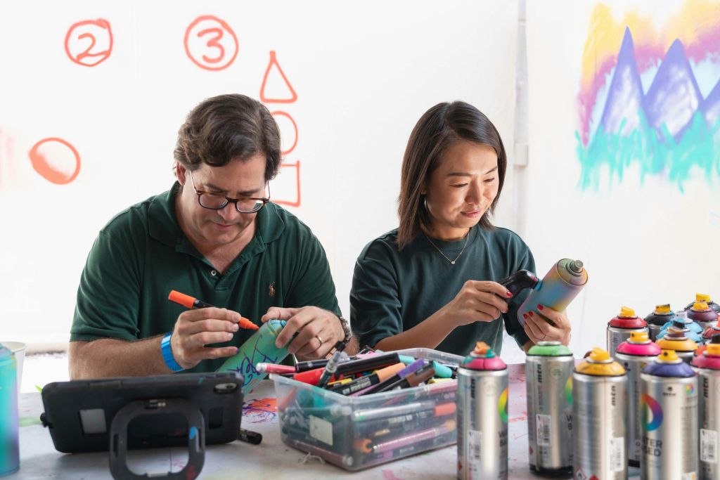 두 사람이 테이블에 앉아 스프레이 페인트와 마커로 작업하고 있습니다. 배경은 다채로운 그래피티와 기하학적 형태를 보여줍니다.