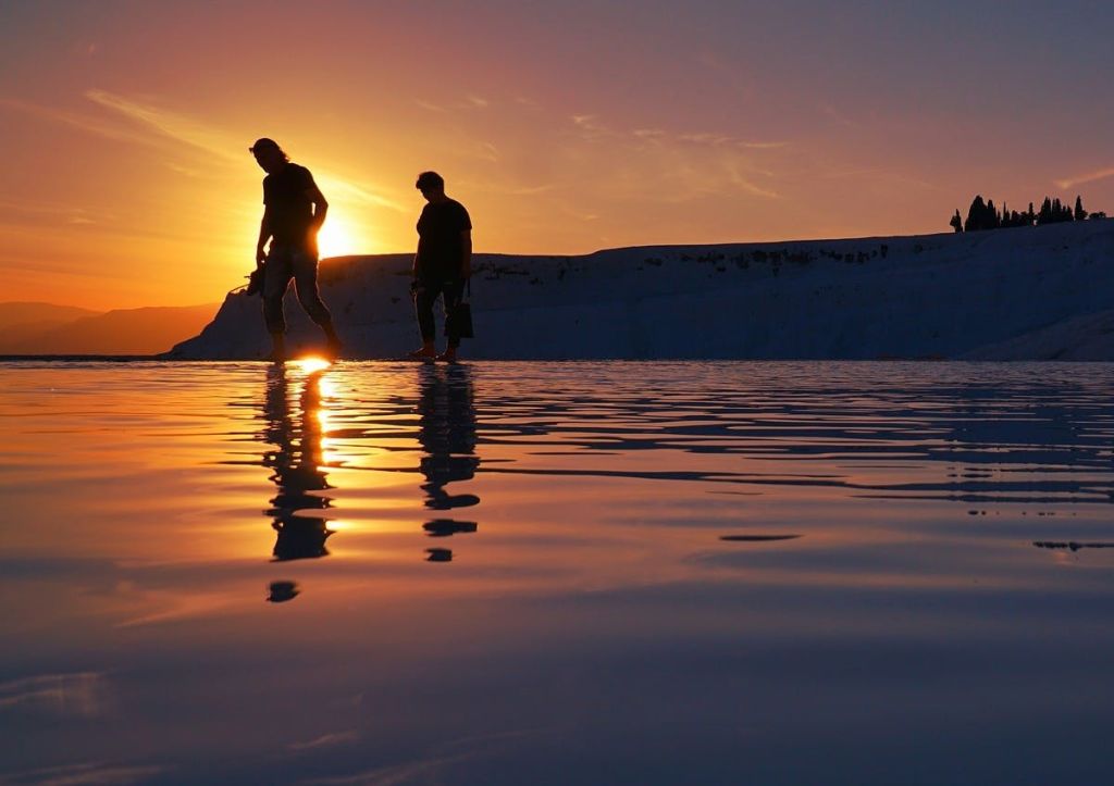 석양을 배경으로 얕은 바다를 거닐고 있는 두 사람.