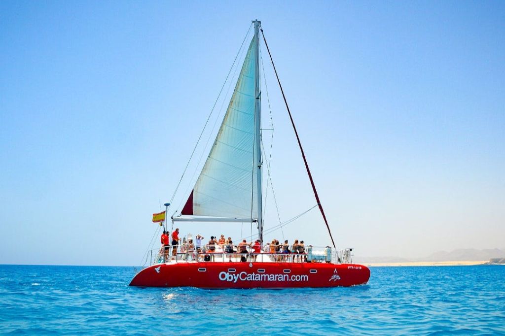 구름 한 점 없는 하늘 아래 맑고 푸른 물 위에 사람들이 돛을 달고 있는 붉은 쌍동선. "ObyCatamaran.com" 이라는 텍스트가 보트에 표시됩니다.