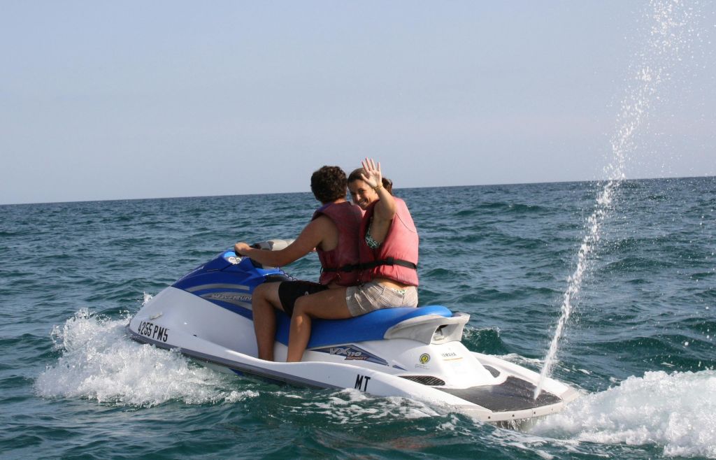 구명조끼를 입은 두 사람이 바다에서 제트스키를 타고 있습니다. 뒤쪽에 있는 사람이 카메라를 향해 유쾌하게 물결칩니다.