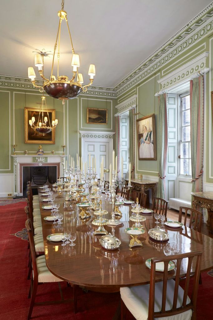 긴 나무 테이블이 있는 우아한 식당으로, 식기와 촛대가 놓여 있습니다. 샹들리에, 그림, 높은 창문이 이 공간을 장식합니다.