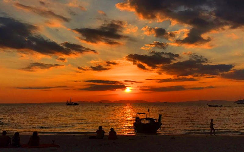 두 사람이 해변에 앉아 화려한 하늘을 배경으로 실루엣의 보트를 타고 바다의 석양을 감상하고 있습니다.