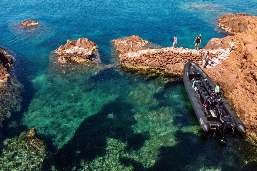 맑고 푸른 물가 바위 부두에 사람들이 서 있고, 근처에 고무 보트가 정박해 있습니다.