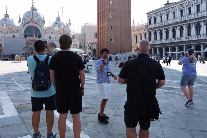 높은 벽돌 타워와 화려한 건물이 있는 유서 깊은 광장에서 투어 가이드가 세 명의 남자와 이야기를 나눕니다.