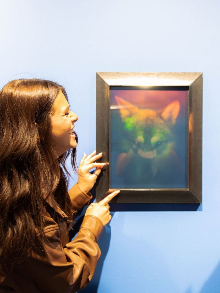 푸른 벽에 걸린 여우의 홀로그램 액자 그림을 가리키며 미소를 짓고 있는 여성.