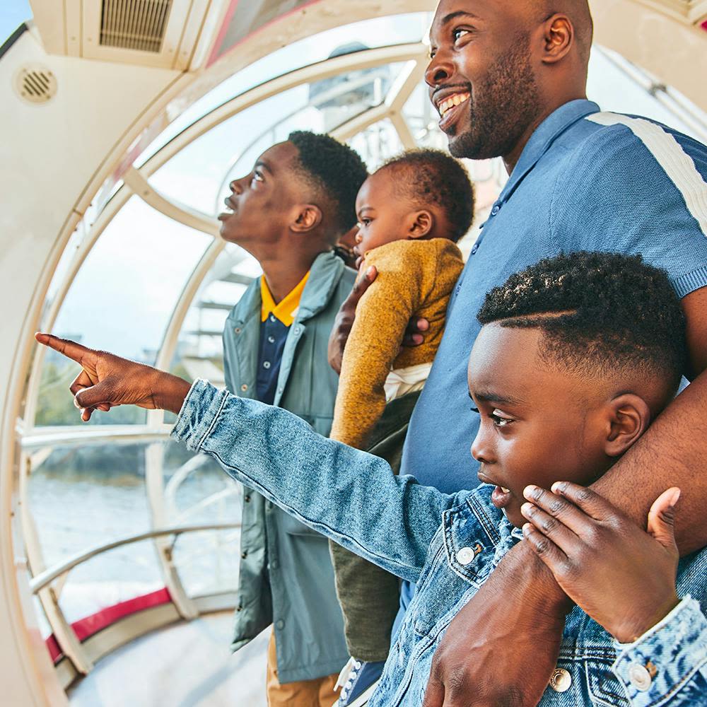 유리벽 구조물 안에서 두 아이와 함께 서 있는 동안 웃는 남자가 아기를 안고 있습니다. 한 아이가 바깥을 가리키고 있습니다.