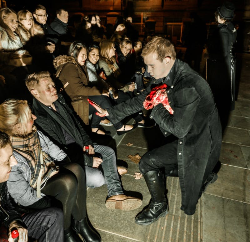 어두운 색의 코트를 입은 남성이 가짜 장기로 보이는 것을 들고 앉은 관객을 위해 공연하고, 다른 사람이 배경에 서 있습니다.