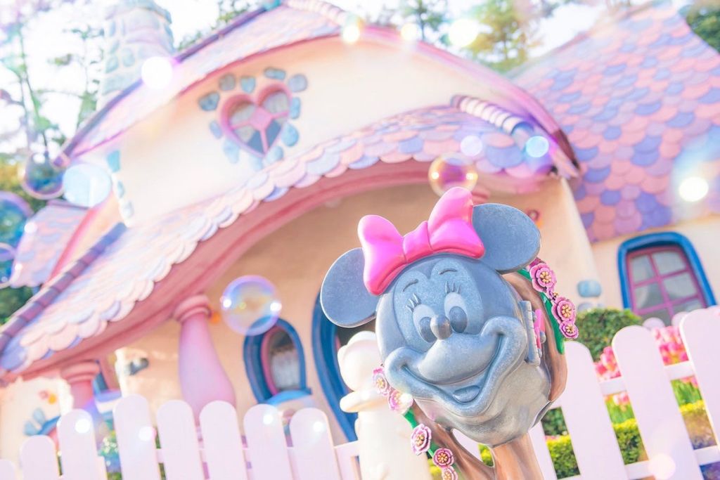 하트 모양의 장식과 파스텔 색상의 기발한 집 앞에 분홍색 활이 있는 미니 마우스 동상입니다.