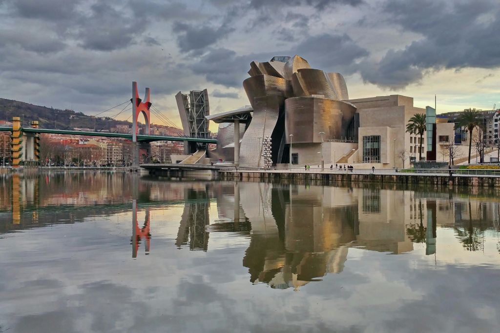 붉은 다리와 흐린 하늘을 배경으로 잔잔한 물에 반사되는 현대적인 금속 건물. 수변에 보행자가 많지 않습니다.