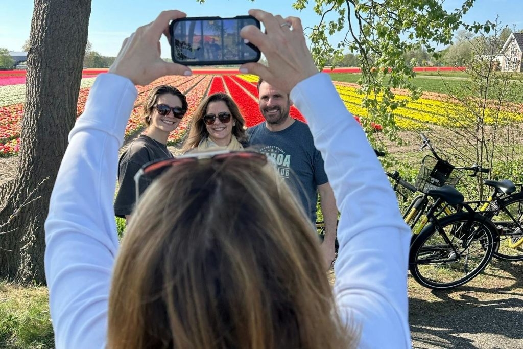 화려한 꽃밭 앞에 웃고 있는 세 사람의 사진을 촬영합니다. 배경에 나무와 가지가 있습니다.