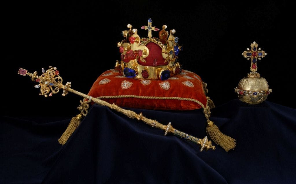 어두운 배경에 금색으로 포인트를 준 레드 벨벳 쿠션 위에 보석으로 장식된 왕관과 홀이 놓여 있습니다.