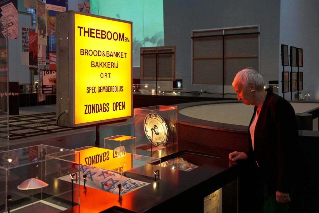 한 노인이 조명 디스플레이 케이스와 박물관 같은 분위기에서 "THEEBOOM BV." 라고 쓰인 밝은 노란색 베이커리 간판을 바라보고 있습니다.