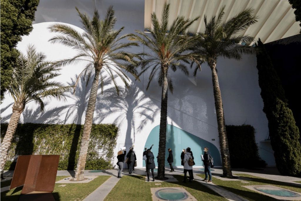 사람들은 커다란 야자수 아래에서 곡선형의 청록색 입구가 있는 현대적인 건물에 그림자를 드리우며 산책을 합니다.