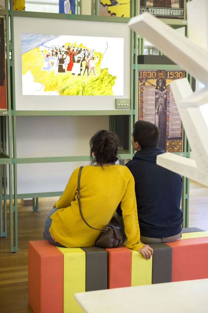 두 사람이 형형색색의 벤치에 앉아 지도와 일러스트레이션이 있는 디스플레이 화면을 바라보고 있습니다. 인근에 역사적인 포스터가 있습니다.