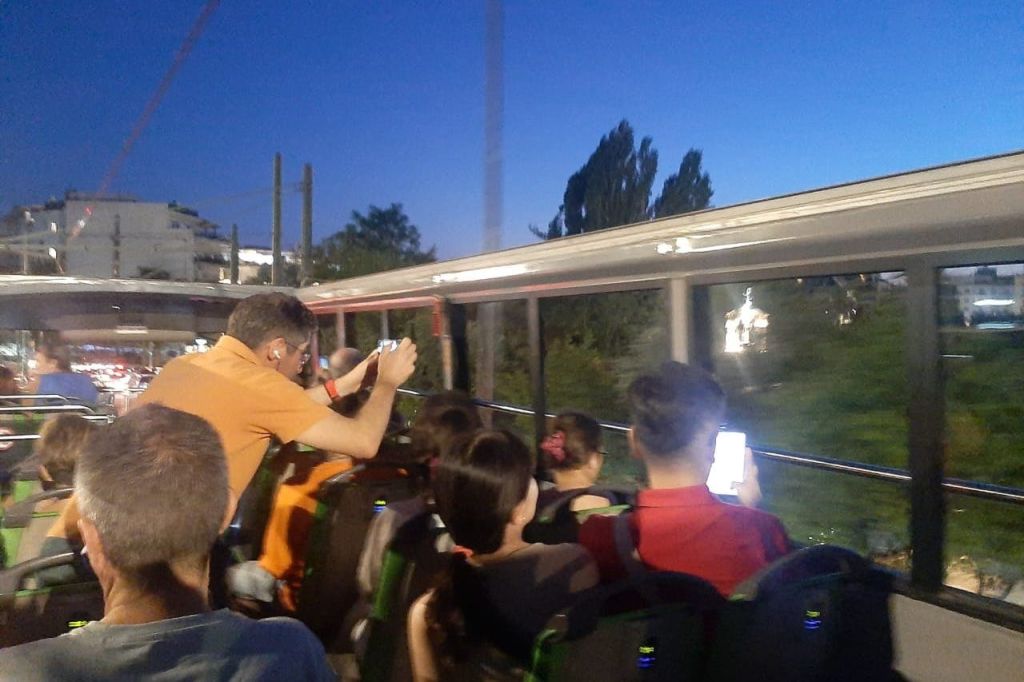 해질 무렵 투어 버스의 상부 갑판에 앉아 있는 성인과 어린이의 모습과 주황색 셔츠를 입은 한 남성이 사진을 찍고 있습니다.