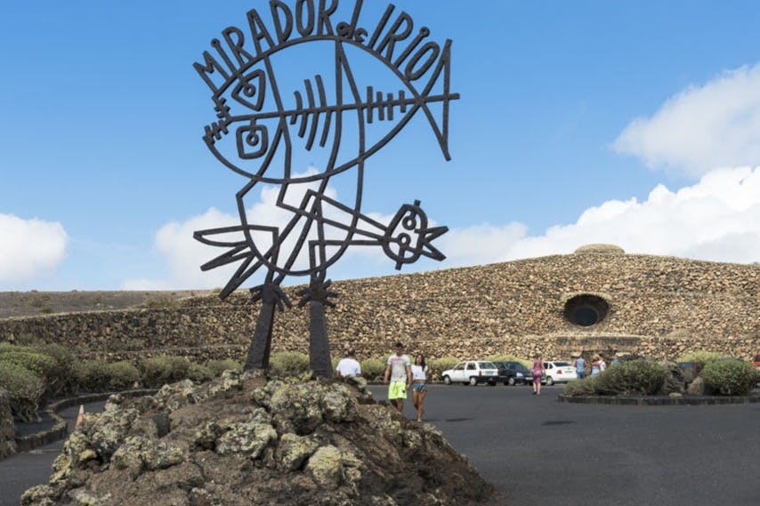 원형 창문이 있는 석조 건물 앞에 "미라도르 델 리오" 라고 적힌 금속 조각상이 서 있습니다. 인근에 사람과 주차된 차량이 있습니다.