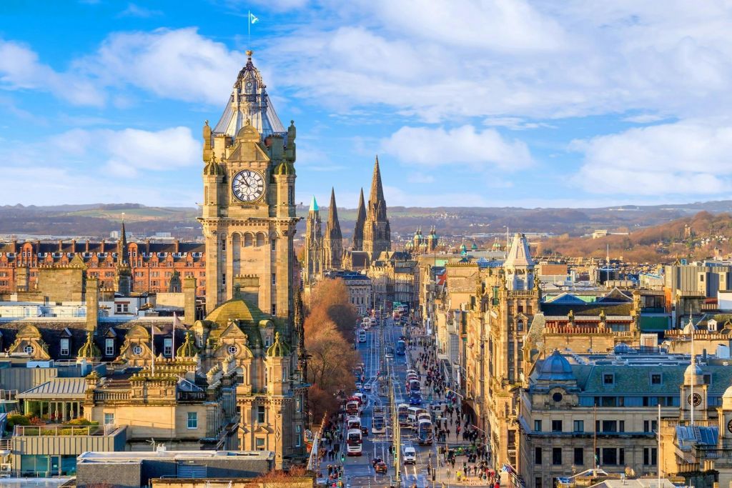 부분적으로 흐린 하늘 아래 시계탑, 첨탑, 자동차와 버스가 있는 번화한 거리가 있는 도시 풍경.