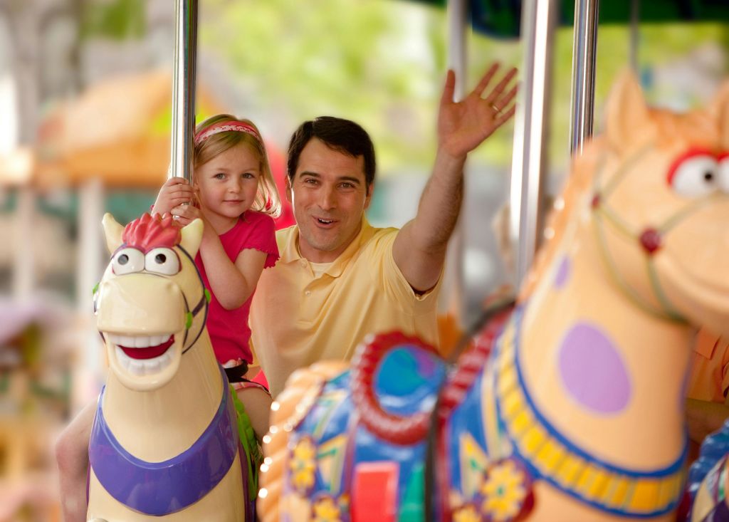 남자와 어린 소녀가 회전목마를 타고 있습니다. 소녀가 기둥을 붙잡고 있는 동안 남자는 미소를 지으며 손을 흔들었다. 밝고 다채로운 요소가 둘러싸고 있습니다.