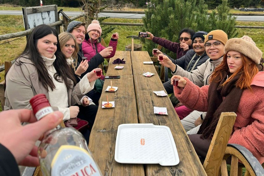 4명이 야외 나무 테이블에 앉아 안경을 들고 있습니다. 병과 트레이가 전면에 있으며, 배경에는 추가 인원이 있습니다.