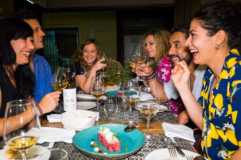 음식 요리와 와인 한 병이 놓인 테이블 주위에서 와인잔을 들고 웃으며 건배하는 네 사람.