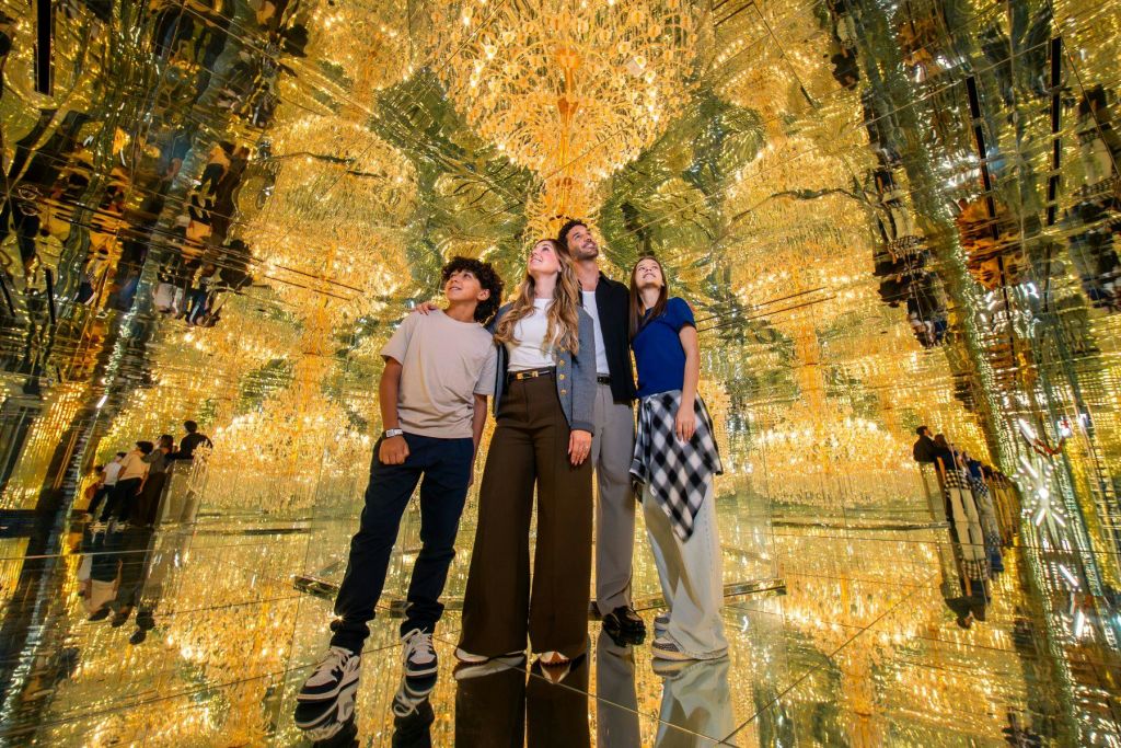 황금빛 샹들리에 같은 빛 반사가 가득한 거울 방에 네 명이 함께 서 있습니다.