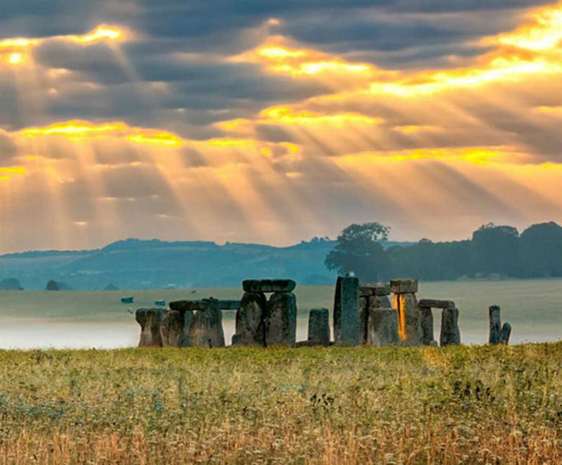햇살이 구름을 뚫고 들어오는 일출의 스톤헨지는 고대 돌과 잔디가 우거진 풍경을 비춥니다.