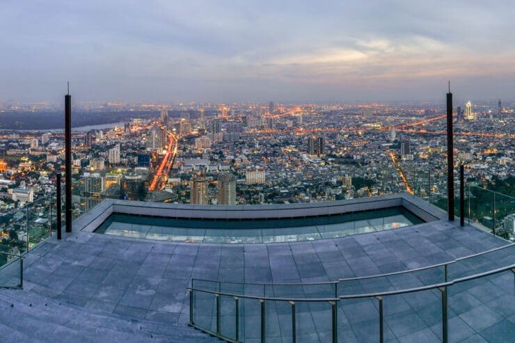 유리 난간이 있는 옥상에서 바라보는 도시 풍경, 해질 무렵에 조명이 켜진 거리가 있는 도심 지역이 내려다보입니다.