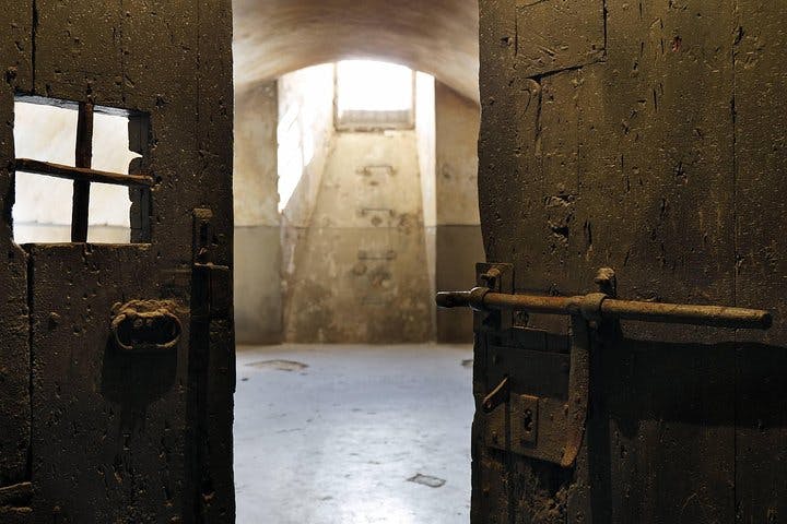 아치형 천장과 작고 막힌 창문이 있는 조명이 희미한 빈 방으로, 탁 트인 녹슨 금속 문이 보입니다.