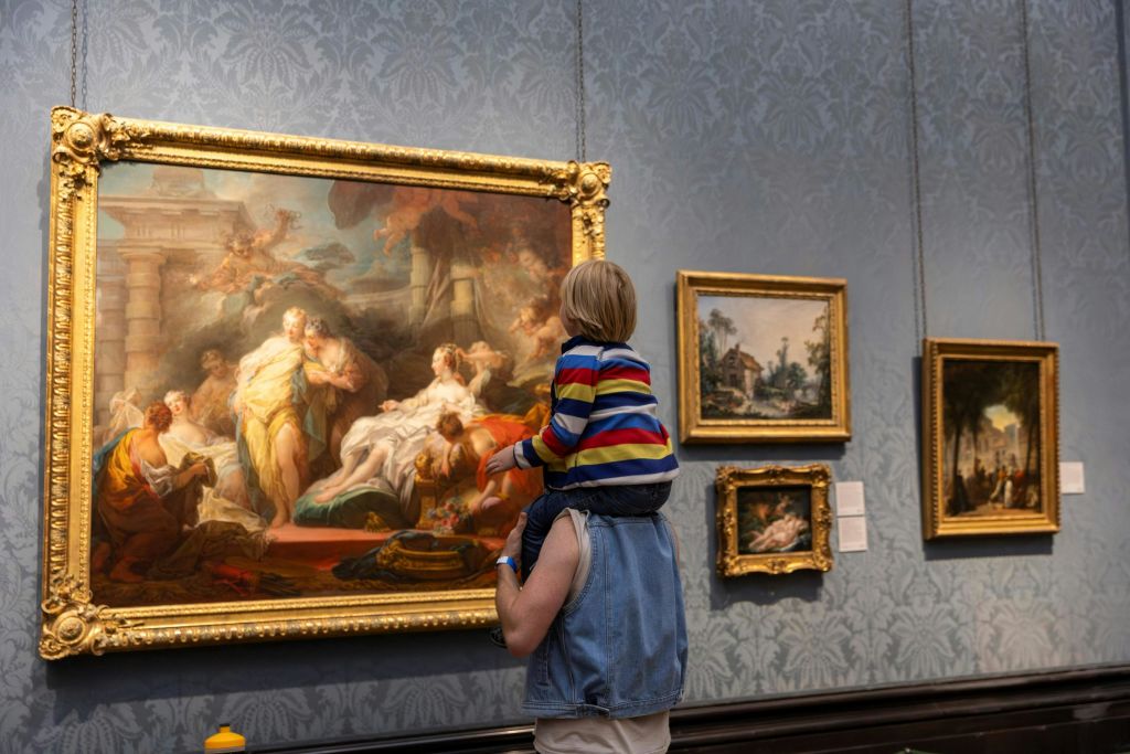 어른의 어깨에 앉아 미술관에서 크고 화려한 그림을 보고 있는 어린이로, 벽에 다른 액자의 예술품이 있습니다.