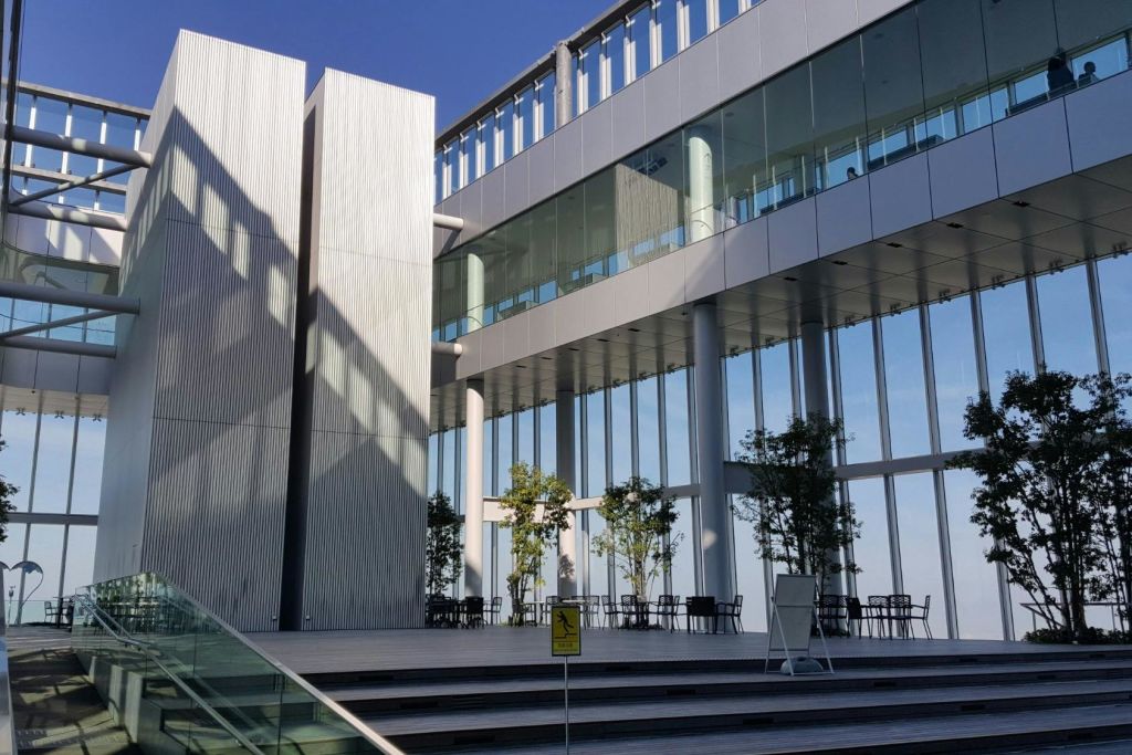 커다란 유리창, 화분의 나무, 야외 좌석이 있는 현대적인 건물. 계단과 주의 표지판이 전면에 있습니다.