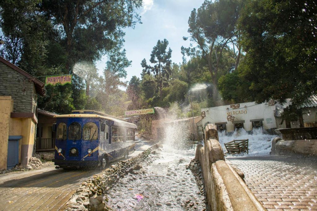 파열된 파이프에서 물이 튀고 나무와 건물로 둘러싸인 파란색 트램이 지나갑니다.