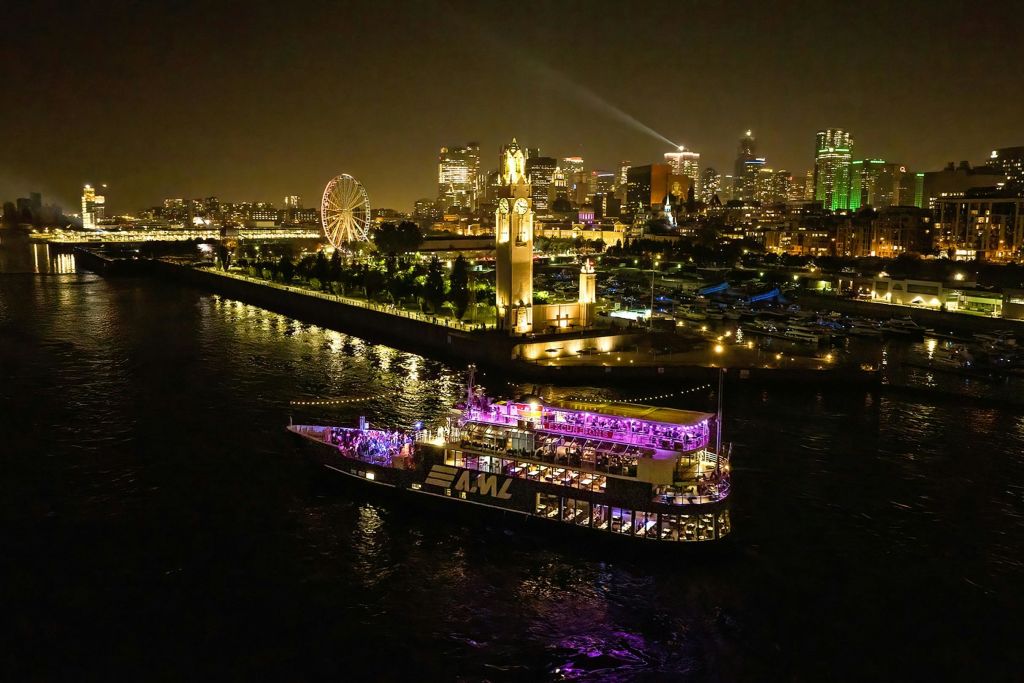 도시의 스카이라인과 조명이 켜진 시계탑을 배경으로 밤에 강에서 환하게 조명이 켜진 보트.