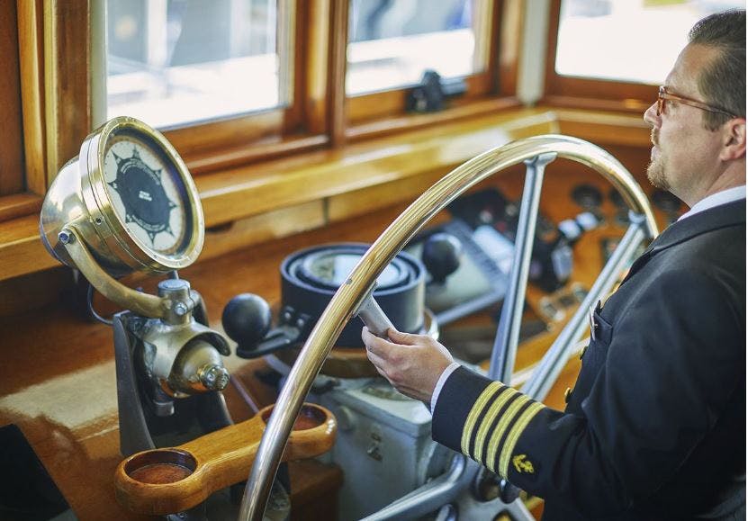선장의 유니폼을 입은 남성이 선박의 다리 안에 있는 큰 바퀴를 사용하여 배를 조종합니다.