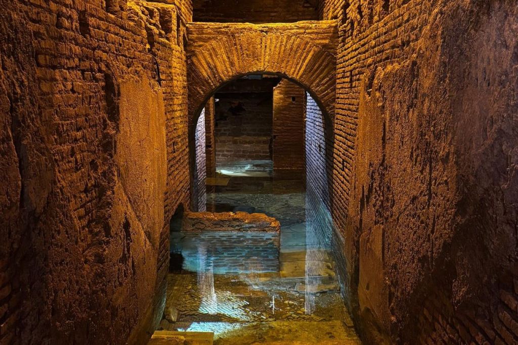 아치형 출입구가 있는 고대 벽돌 복도로, 부분적으로 물에 잠기고 노란색과 파란색 조명으로 조명이 켜져 있습니다.