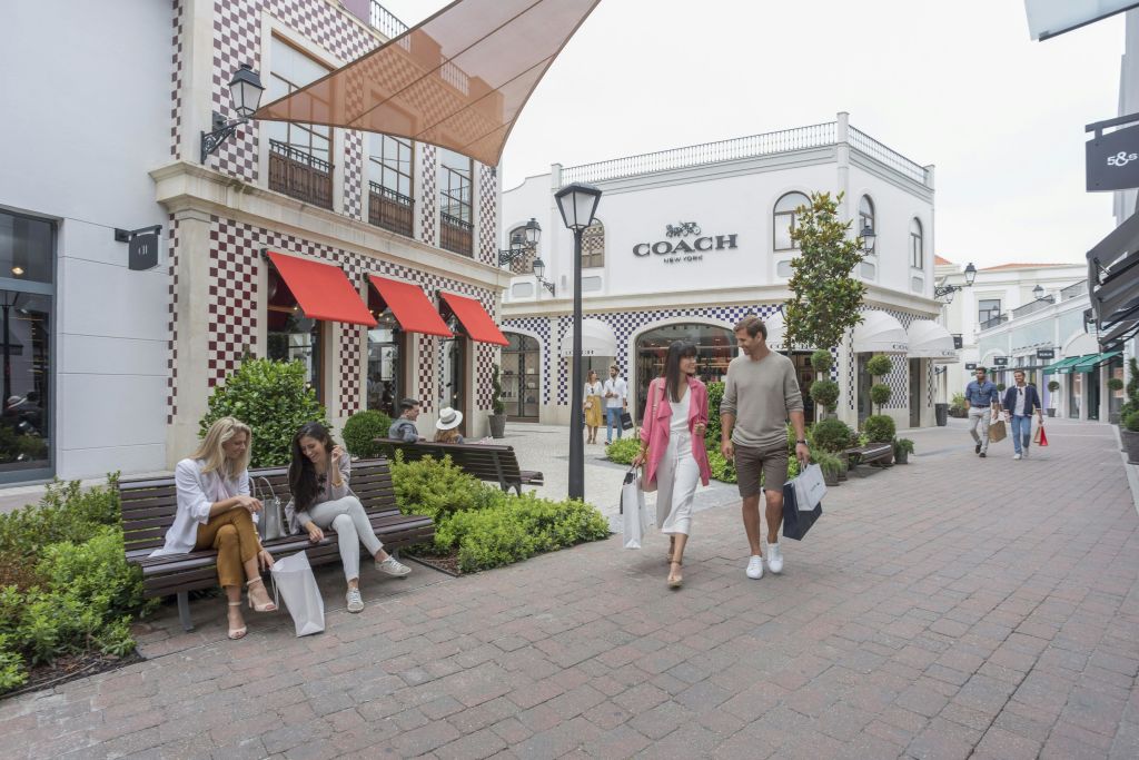 벤치와 녹지로 둘러싸인 Coach 매장을 배경으로 야외 광장에서 걷고 쇼핑하는 사람들.