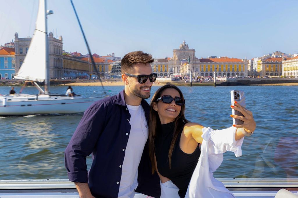 유서 깊은 건물과 요트를 배경으로 한 도시 풍경이 펼쳐지는 바닷가에서 웃고 있는 커플이 셀카를 찍고 있습니다.