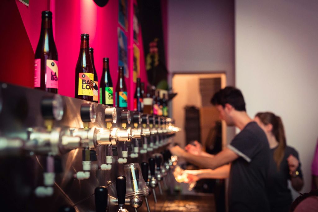 조명이 희미한 바 위에 놓인 선반에 다양한 색상의 병이 놓인 맥주 탭 벽에서 한 남성이 음료를 쏟아내고 있습니다.