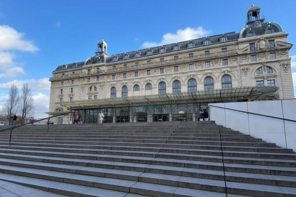 푸른 하늘 아래 넓은 계단 아래에서 바라보는 입구의 유리 캐노피가 있는 웅장한 역사적인 건물입니다.