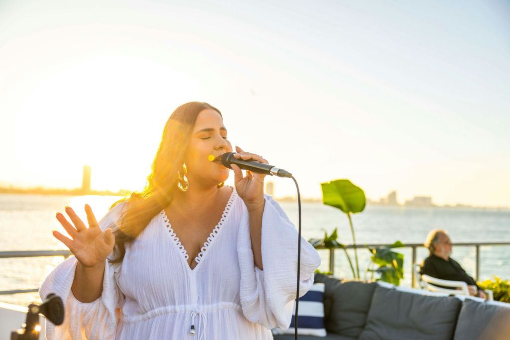 해 질 녘, 물과 멀리 떨어진 건물을 배경으로 한 여성이 야외에서 마이크를 잡고 노래하고 있습니다. 노인이 근처에 앉아 있습니다.