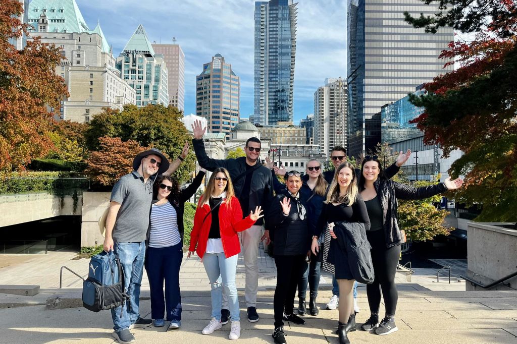 높은 건물과 나무, 맑고 푸른 하늘을 배경으로 야외에서 포즈를 취하고 미소를 짓고 있는 사람들.